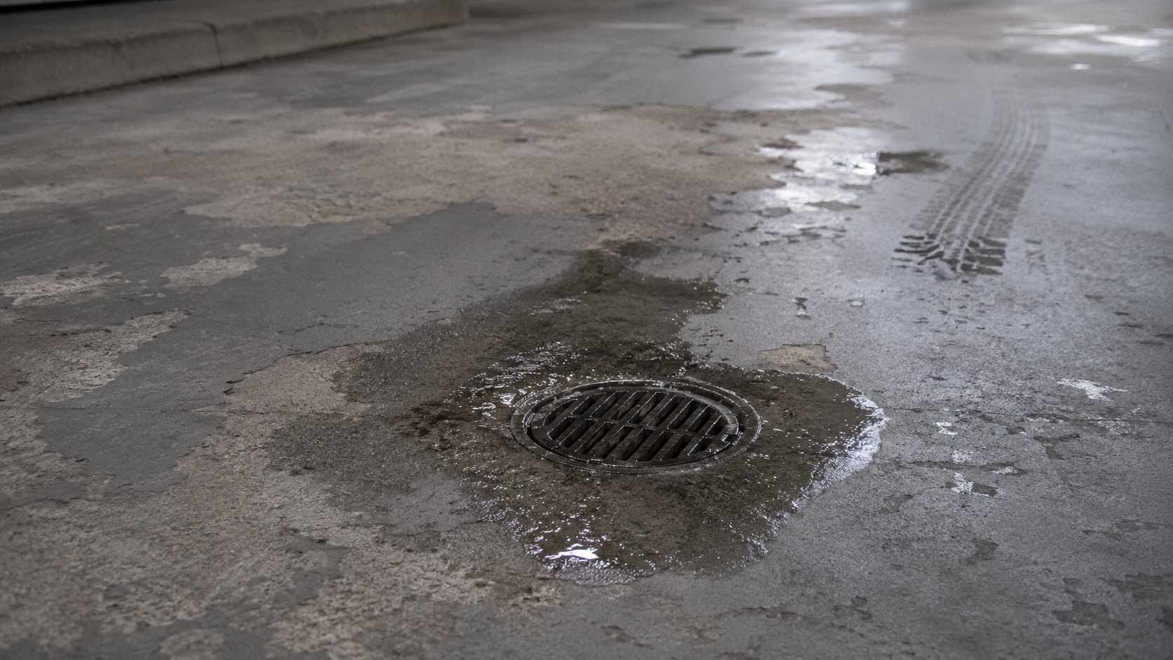 A concrete parking-deck driving surface inside an underground garage showing a worn, cracked traffic-coating membrane with a puddle of water pooling near a floor drain, faint tire marks across the surface, harsh overhead light. No people, no text.
