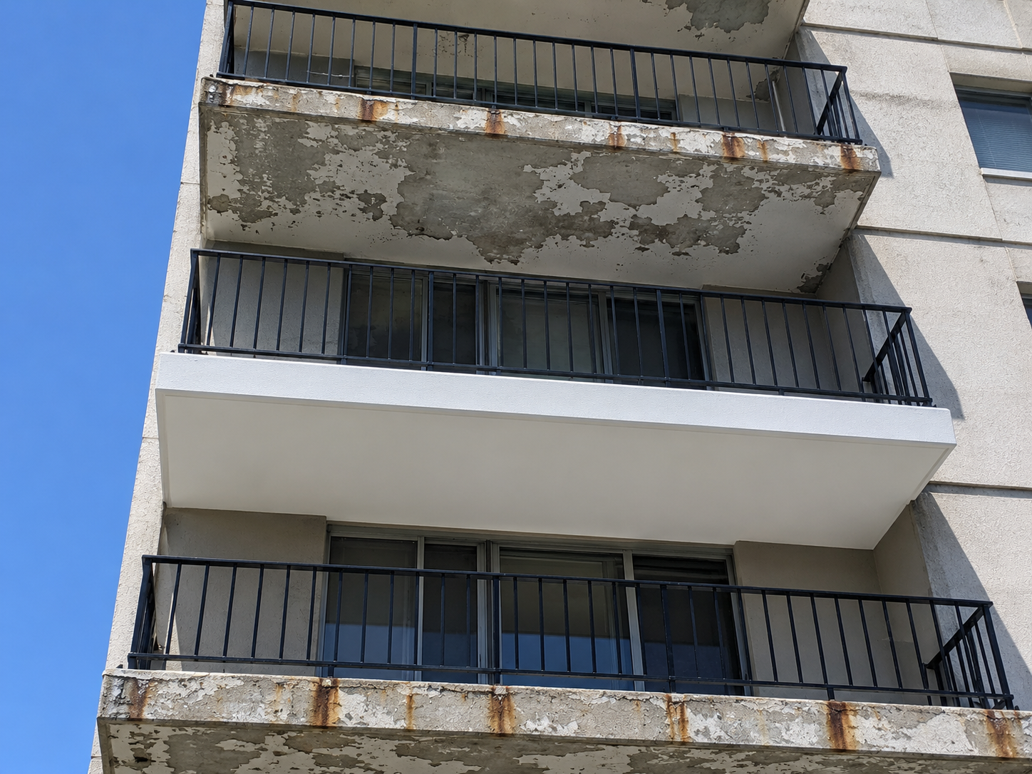 Multi-storey condominium balcony stack in Ottawa with one freshly repainted balcony — clean white ceiling and even railing — contrasting against weathered balconies above and below showing peeling ceiling paint and rust-streaked railings