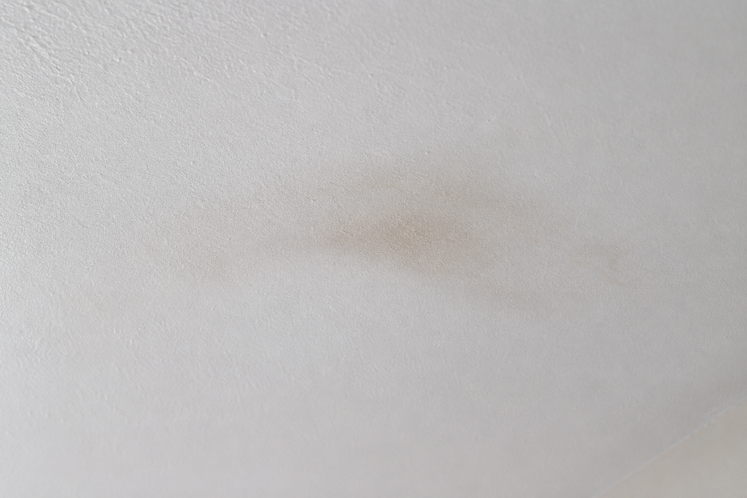 Faint tan water stain bleeding back up through a fresh white paint film on a smooth ceiling under raking light